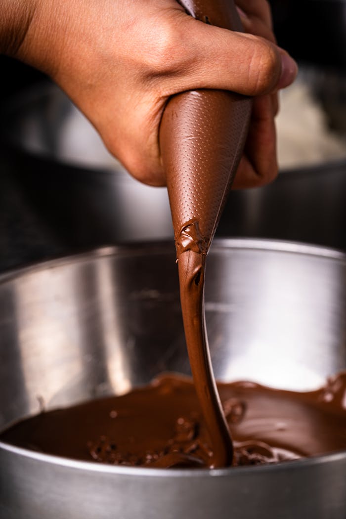 portfolio-img-09 Hand skillfully piping melted chocolate into a stainless steel bowl, focusing on texture and detail.